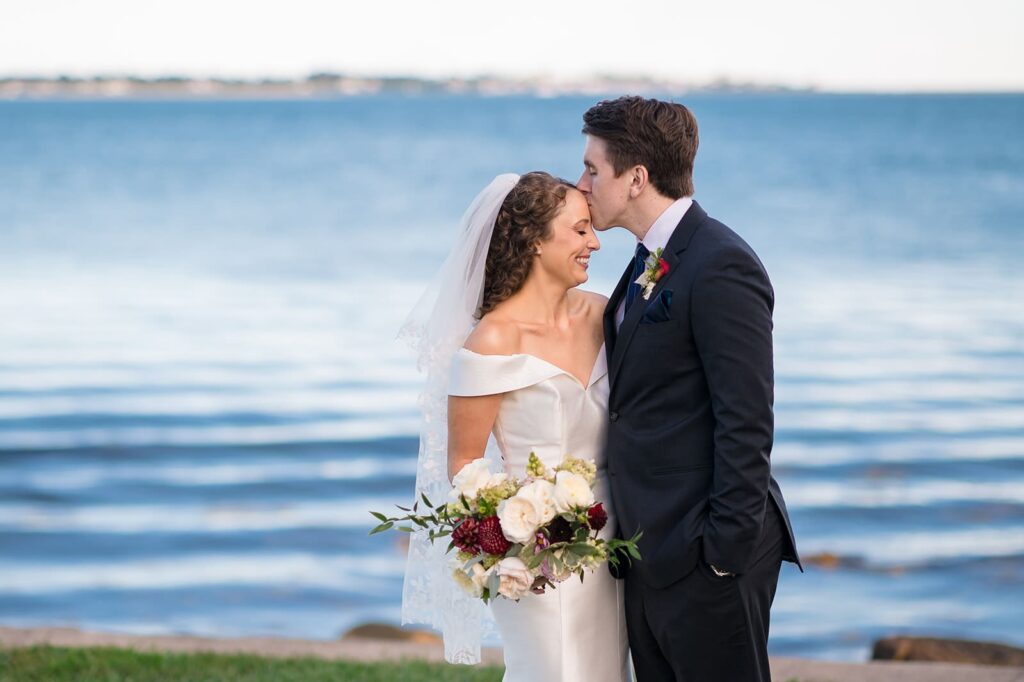 bride and groom stand in front of the ocean with groom kissing bride's forehead at their wedding in stonington ct