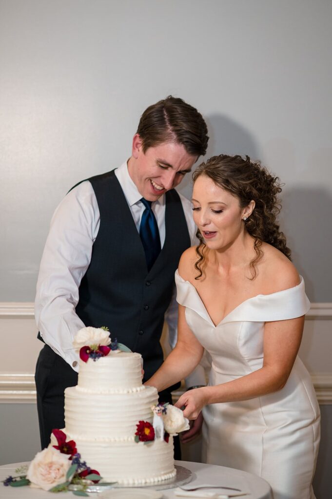 bride and groom smile while cutting their white wedding cake with red and cream flowers