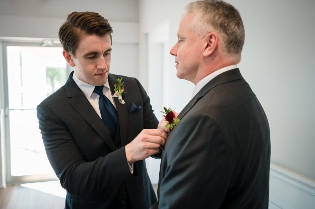Groom wearing a dark suit pins his father's boutonniere onto his jacket.