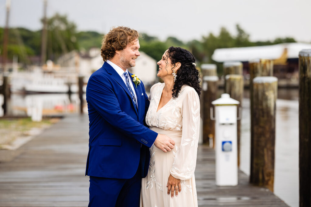 A joyful couple in wedding attire standing on a dock, the groom in a striking blue suit and the bride in a cream dress with detailed sleeves, exchanging smiles by the waterfront