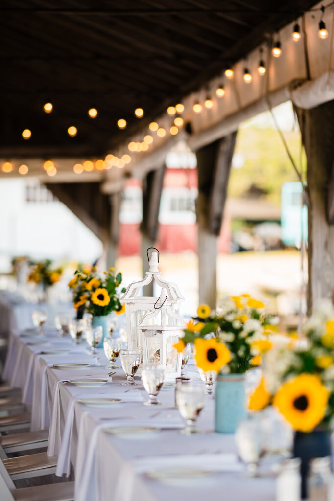 An outdoor wedding table setting under a covered dock adorned with string lights, featuring sunflower centerpieces and a white, elegant table setting, awaiting guests