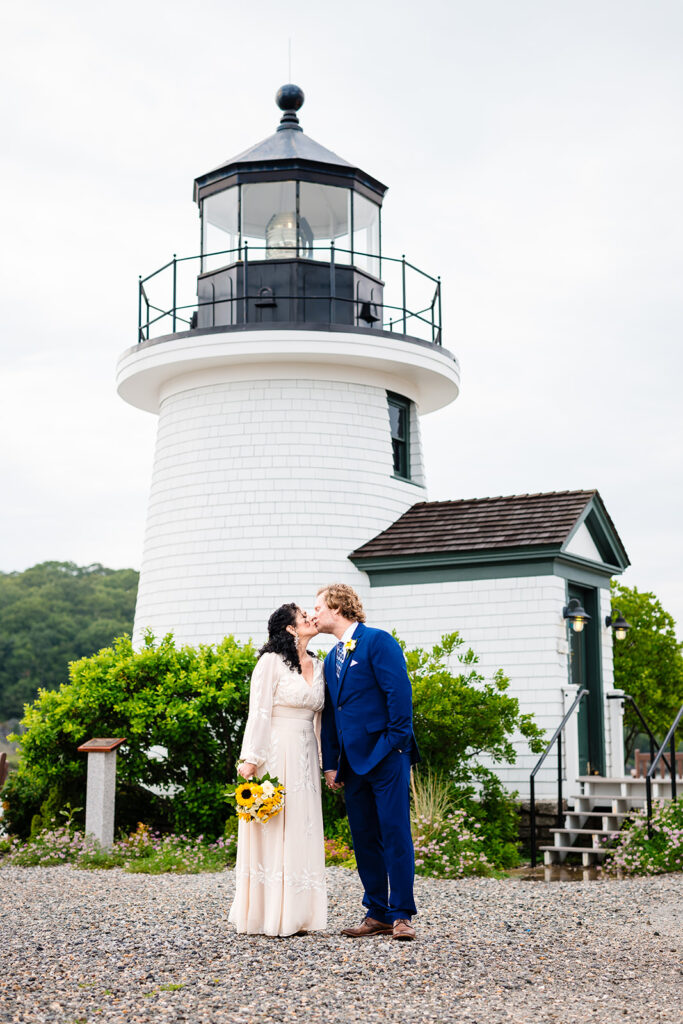 A couple shares a romantic kiss in front of a quaint white lighthouse, the bride holding a bouquet with bright sunflowers, symbolizing a beacon for their journey ahead