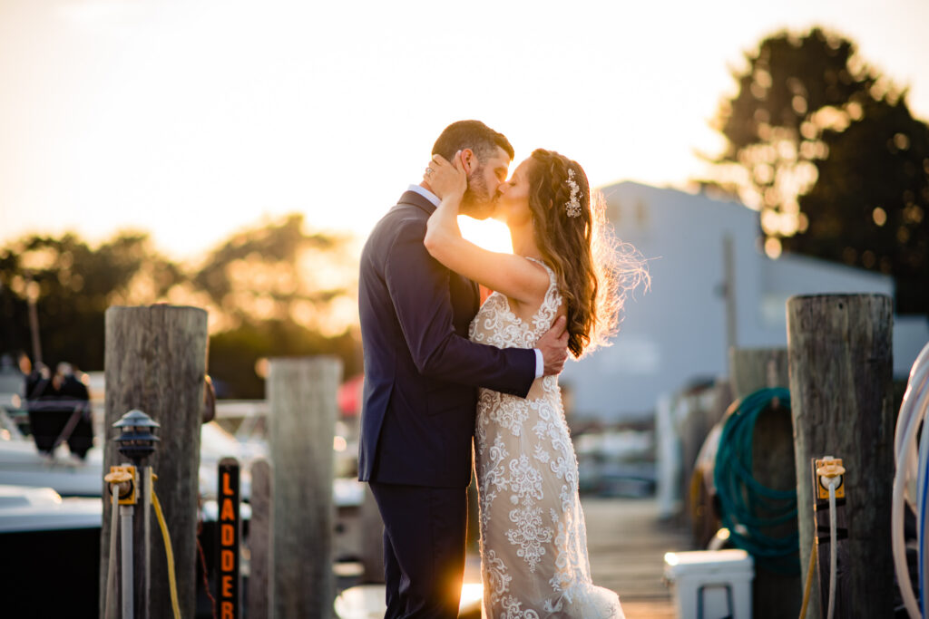 A bride and groom embrace and kiss on a dock at sunset, surrounded by wooden posts, boats, and coiled ropes, with sunlight shining behind them—capturing the romance often found at unique wedding venues in Connecticut.