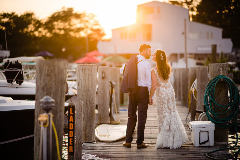 A bride and groom stand on a dock at sunset, facing each other and holding hands, with boats and wooden posts visible in the background—one of the many unique wedding venues in Connecticut that offers a stunning waterfront setting for your special day.