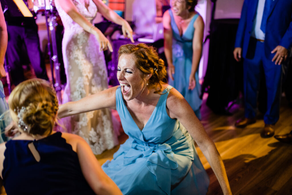 Woman in a blue dress laughs energetically while crouching on a dance floor at an indoor event hosted at one of the unique wedding venues in Connecticut, with other people dancing and smiling in the background.