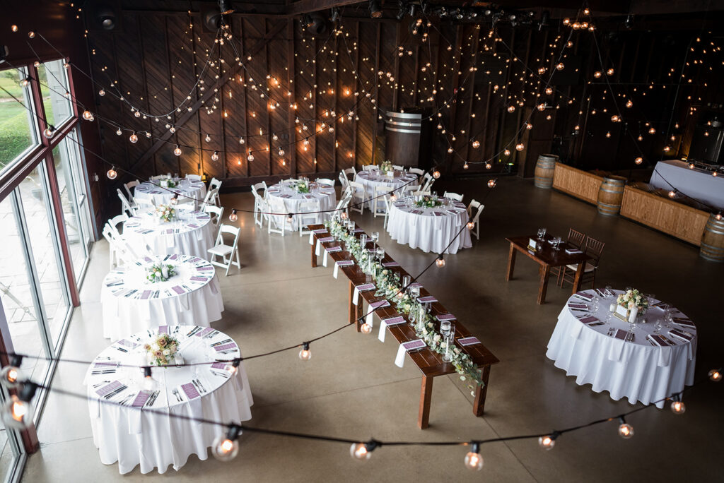 An enchanting wedding reception setup in a rustic barn, illuminated by strands of warm lights crisscrossing the wooden ceiling, with a mix of round and long tables for a cozy atmosphere
