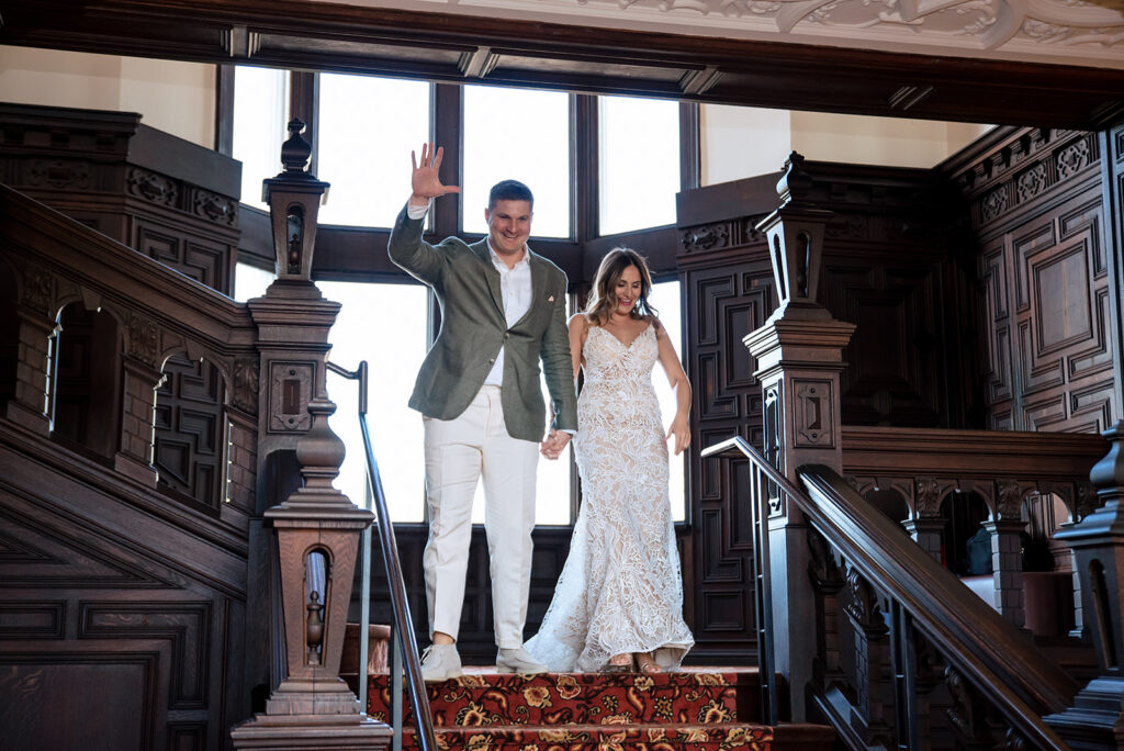 A happy couple descending a grand wooden staircase, the groom in a casual green blazer and white pants, and the bride in a detailed lace wedding dress.