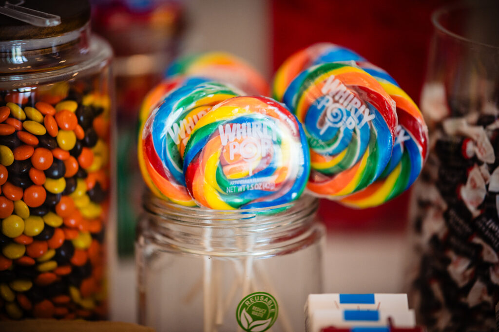 A vibrant Whirly Pop lollipop with rainbow swirls is prominently displayed in a glass jar amidst other candy containers.
