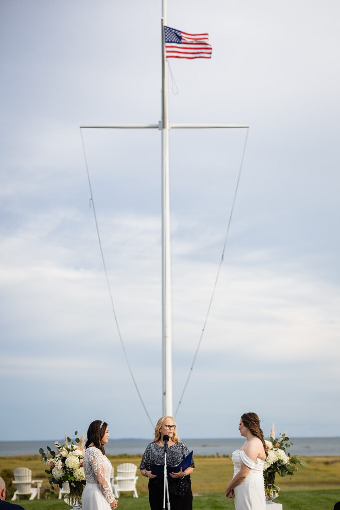 An outdoor wedding scene with two women and an officiant standing before an American flag on a flagpole, with a serene ocean backdrop.