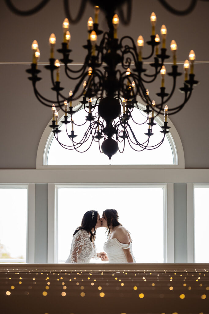 Two brides share a romantic kiss under a black wrought-iron chandelier, silhouetted against a large window with bokeh lights in the foreground.