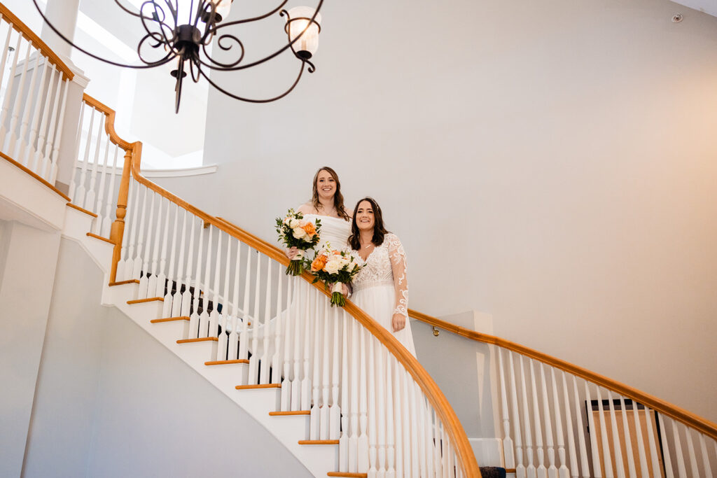 Two brides descend a curved staircase, smiling brightly, with one carrying a bouquet, complemented by a grand chandelier above.