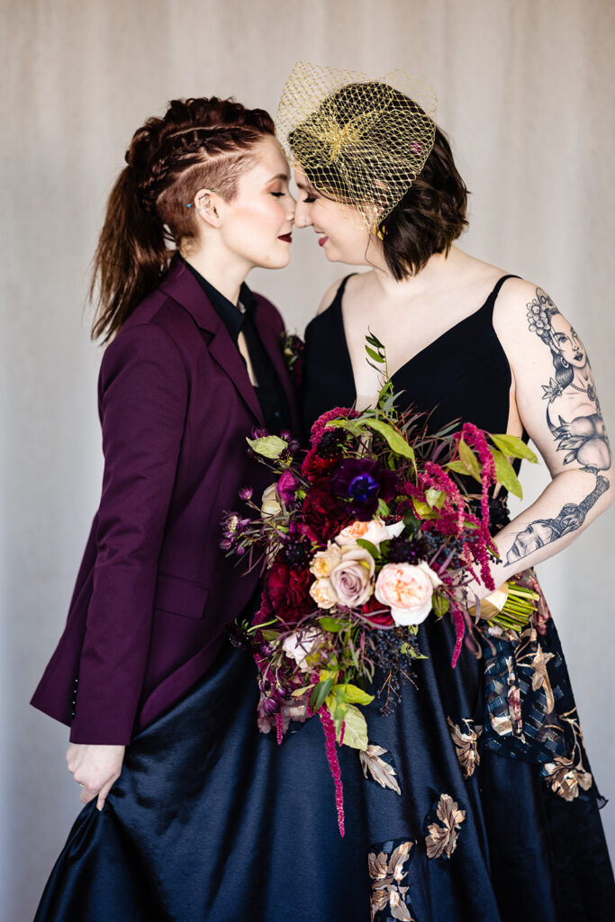 Two women in black and purple gowns kissing in front of a white background.