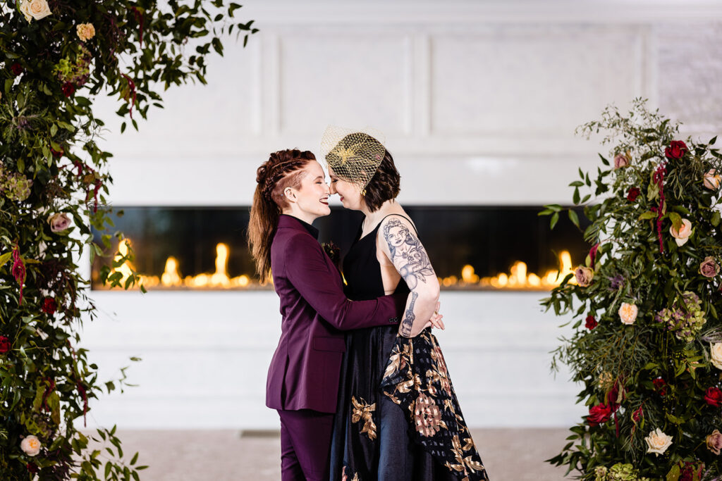 Two brides embrace in front of the fireplace in the ballroom at Grand View Mendon. 