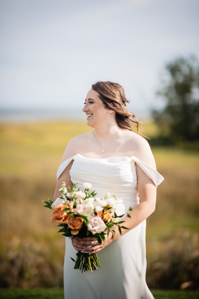 A bride in a white off-shoulder gown holds a bouquet, standing in a field with a broad smile, embodying a sense of joy and anticipation.