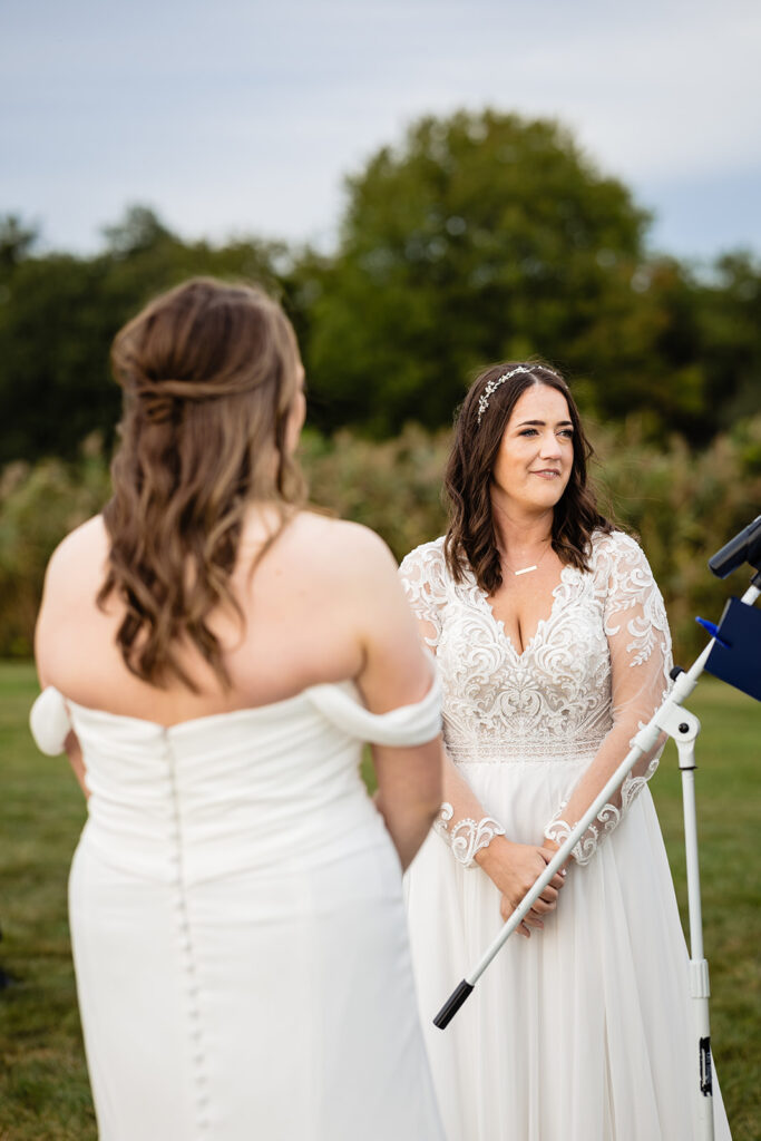 A close-up of two brides at an outdoor ceremony, one in a lace-detailed dress, the other in a simple gown, with a microphone stand between them.
