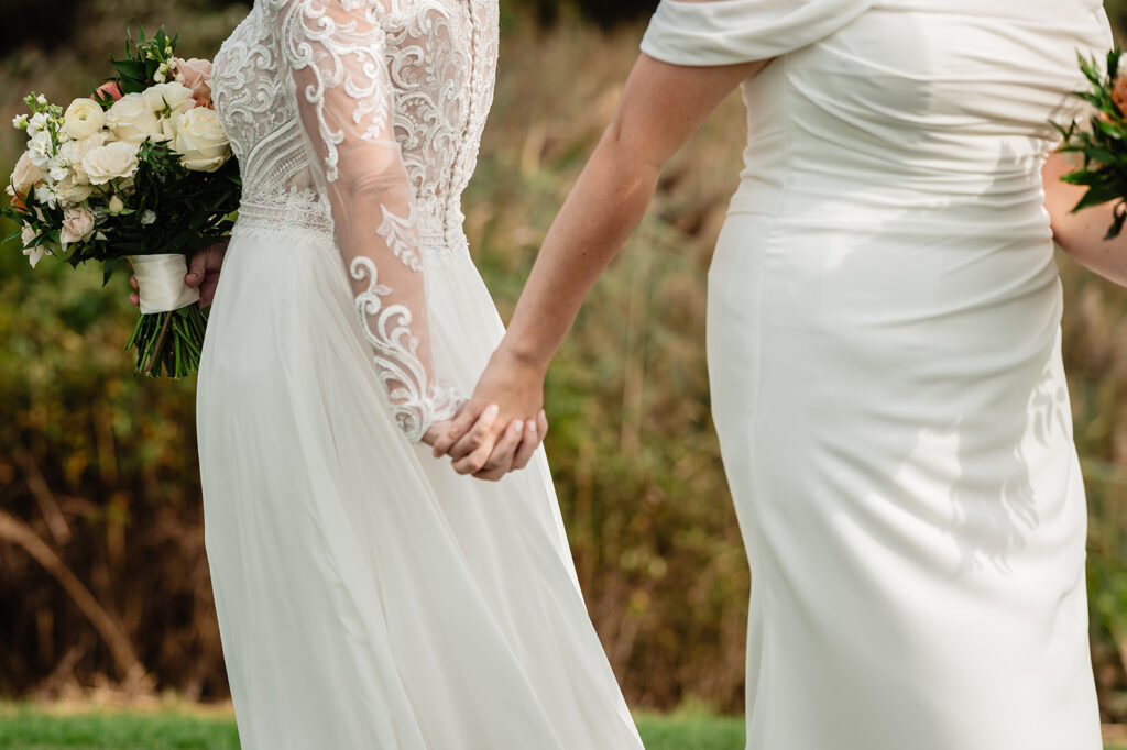 A tender moment as two brides hold hands during their wedding ceremony, showcasing the intricate details of their dresses and the natural beauty of the outdoor setting.