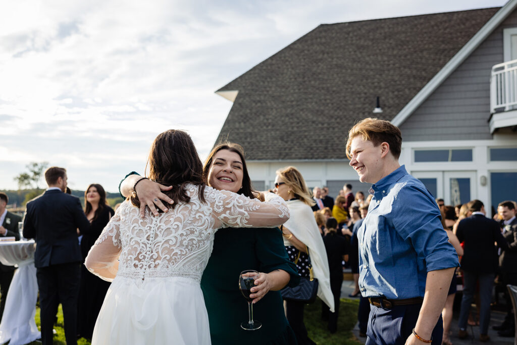 A joyful outdoor wedding scene with a bride in a white lace dress hugging a smiling guest, while a guest in a blue shirt looks on, with other guests mingling in the background.