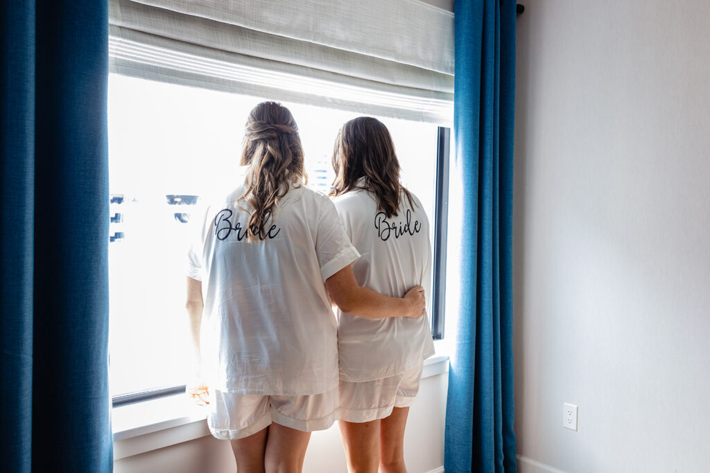 Two women wearing matching white shirts with "Bride" and "Bride" written on the back, standing by a window and looking out, symbolizing the anticipation before a wedding ceremony.