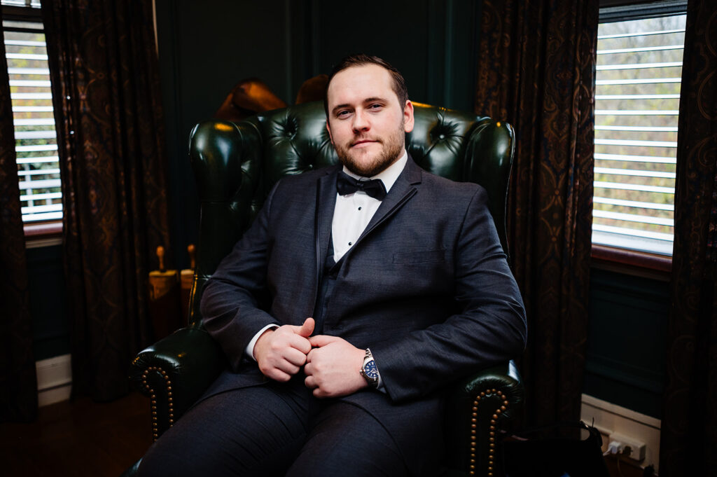 "A man in a dark suit with a bow tie sitting confidently in a vintage green leather armchair, adding a classic touch to the wedding ambiance
