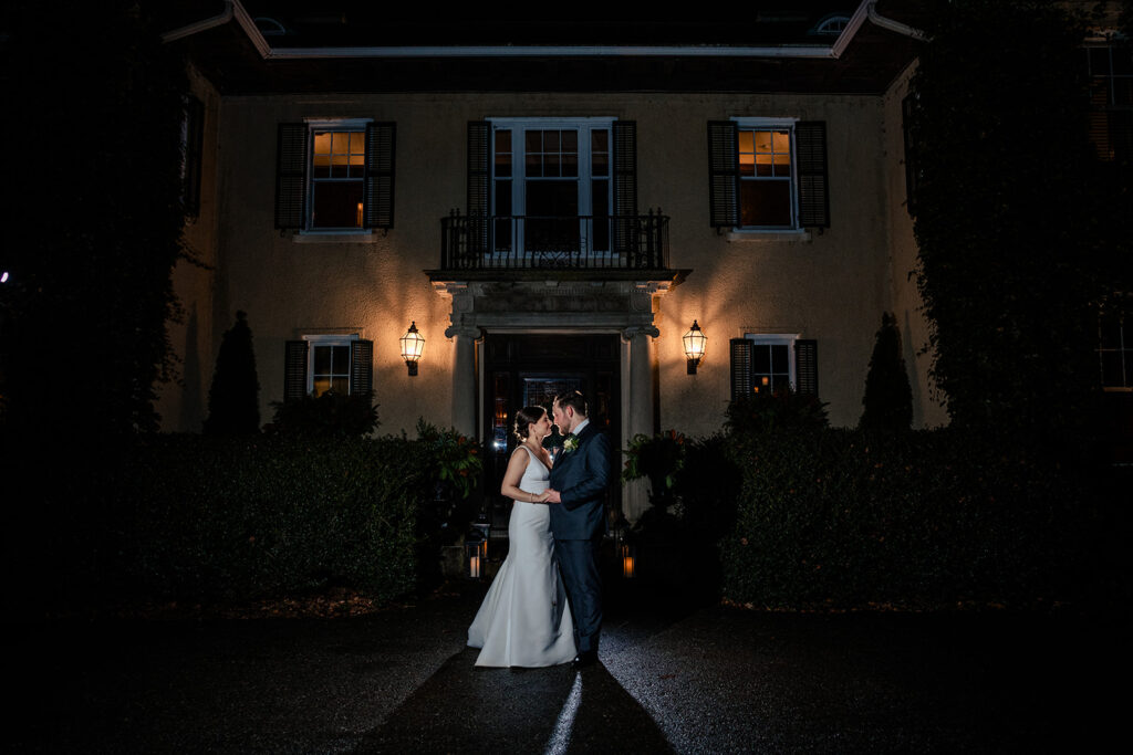 A night shot of a bride and groom sharing an intimate moment in front of a classic manor, beautifully lit to highlight the architecture and the couple's connection