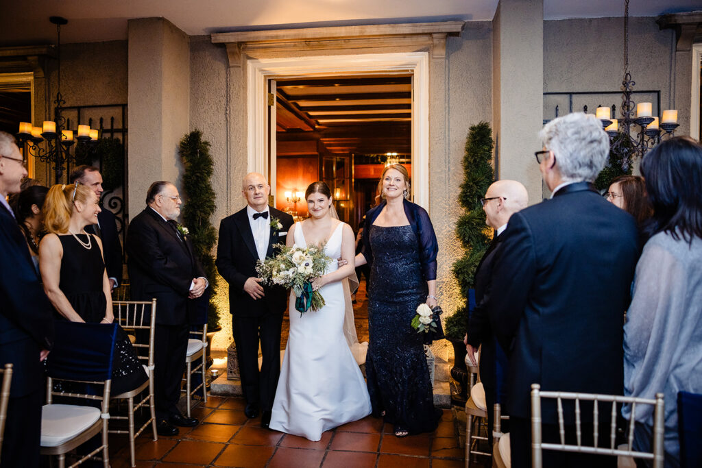 A bride in a simple white gown with a bouquet and an older woman in a dark blue dress smiling as they walk through a doorway, greeted by elegantly dressed guests at a wedding