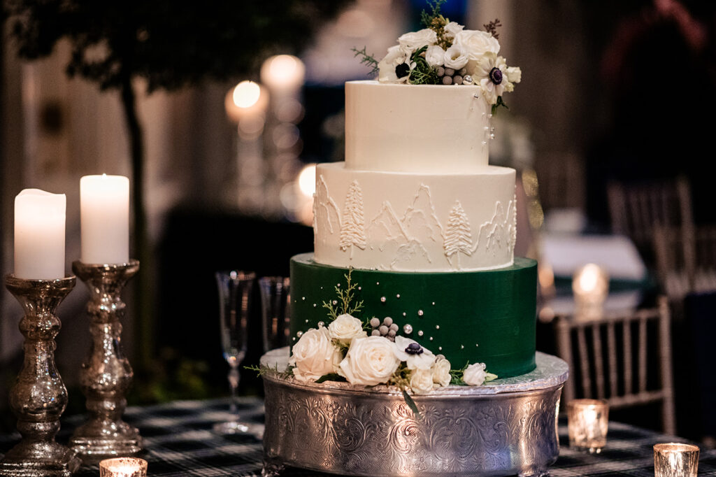 A three-tiered wedding cake with white and green icing, decorated with white flowers and pine tree icing details, set on a silver stand with candles around it, exuding an elegant vibe.