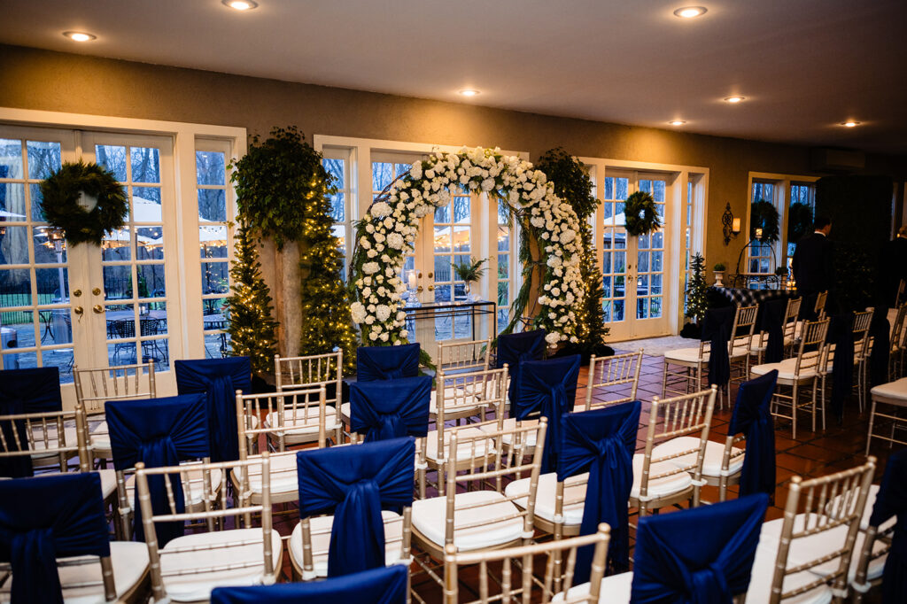 An indoor wedding ceremony venue with white chairs adorned with navy blue sashes, a white floral arch, and strings of lights creating an intimate atmosphere.