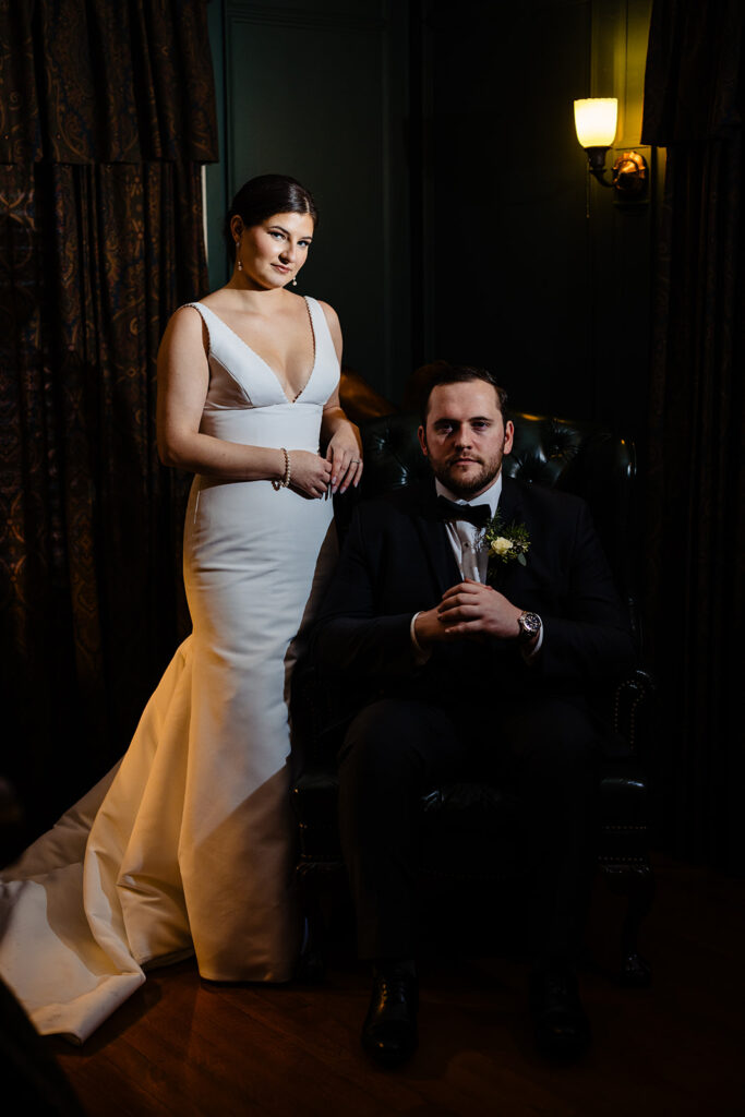 A poised bride in a white dress standing beside a seated groom in a black tuxedo, both looking intently towards the camera, against a dark, luxurious backdrop.