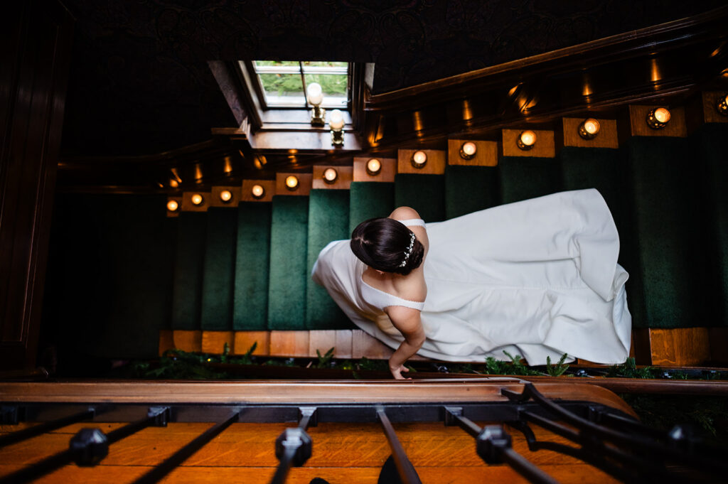 An overhead view of a bride in a flowing white dress ascending a richly carpeted staircase, capturing a moment of bridal anticipation
