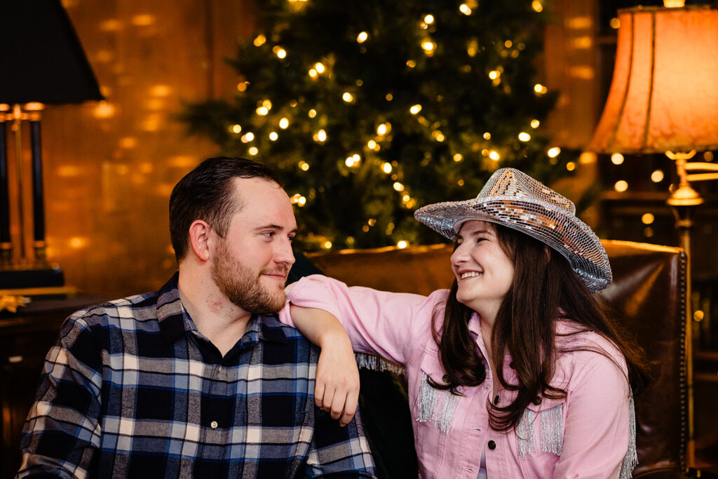 A cozy moment where a couple is laughing together on a sofa, with the woman wearing a cowboy hat, against a backdrop of a twinkling Christmas tree