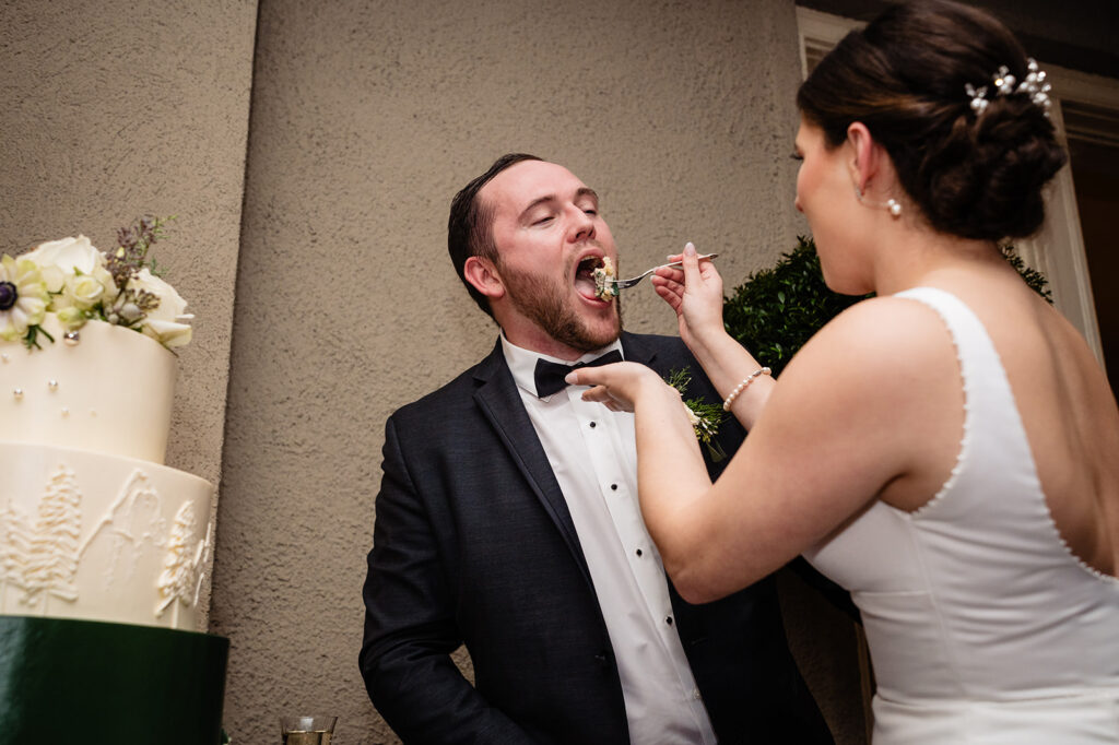 A playful moment where a bride feeds the groom a piece of wedding cake, both dressed in formal wedding attire, with guests in the background