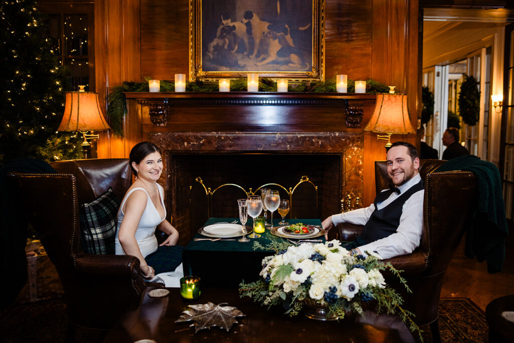 A charming dinner scene with the bride and groom seated in leather armchairs by the fireplace, smiling at each other, surrounded by festive decorations and a beautiful floral arrangement
