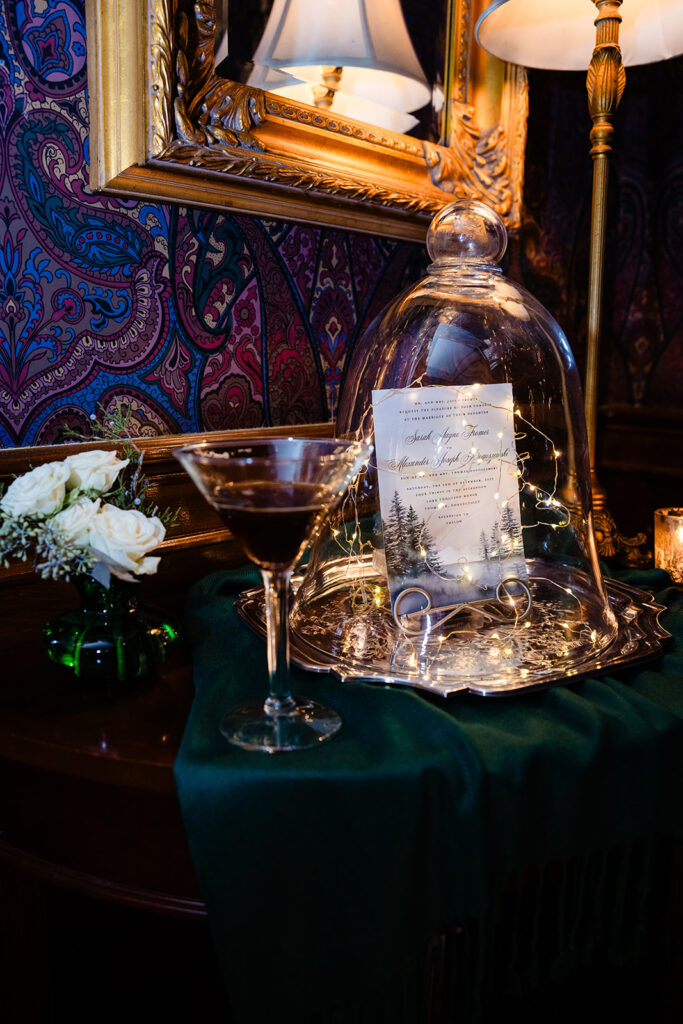 A sophisticated wedding invitation under a glass dome, accompanied by a cocktail and floral decor, with rich fabric in the background, invoking a sense of timeless elegance