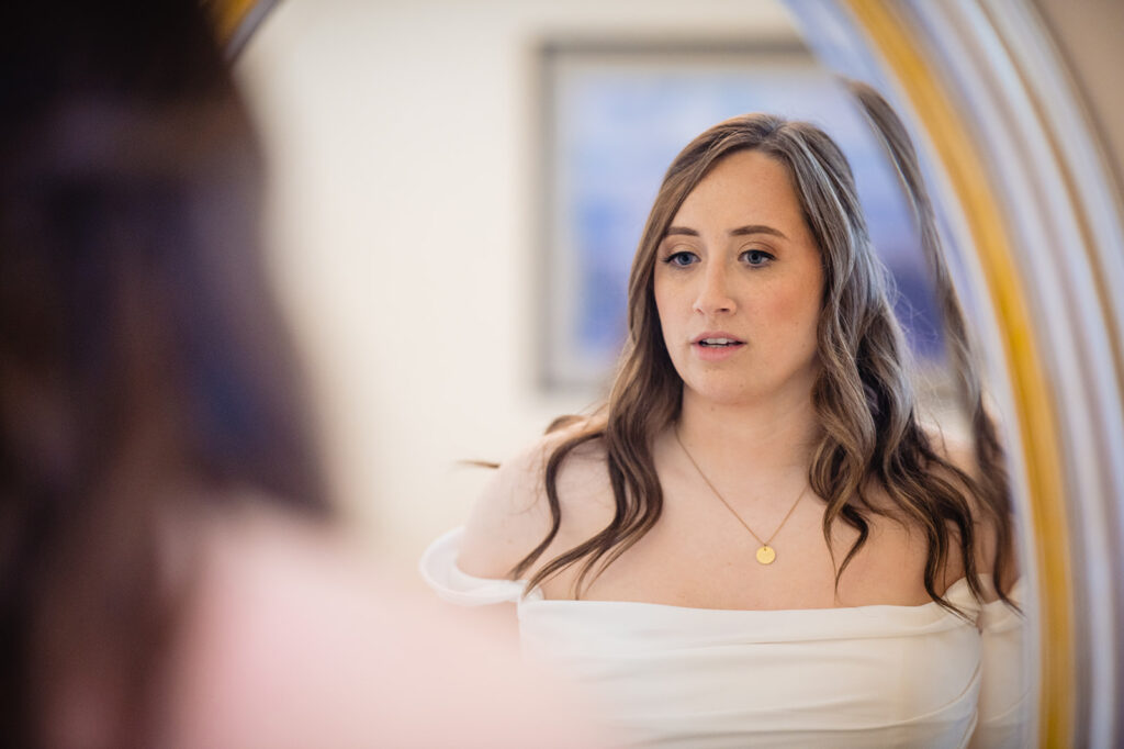 A bride reflected in an oval mirror, looking contemplative as she prepares for her wedding, with another person in the reflection assisting her.