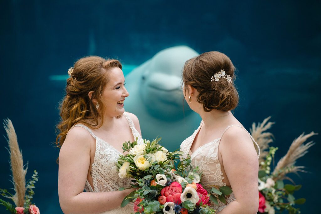 Two bridesmaids in lacy dresses sharing a joyful moment in front of a serene beluga whale exhibit, highlighting a unique wedding backdrop