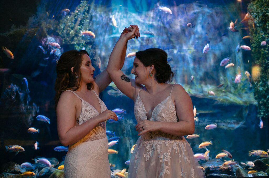 Two brides in white lace dresses dance in front of a colorful aquarium