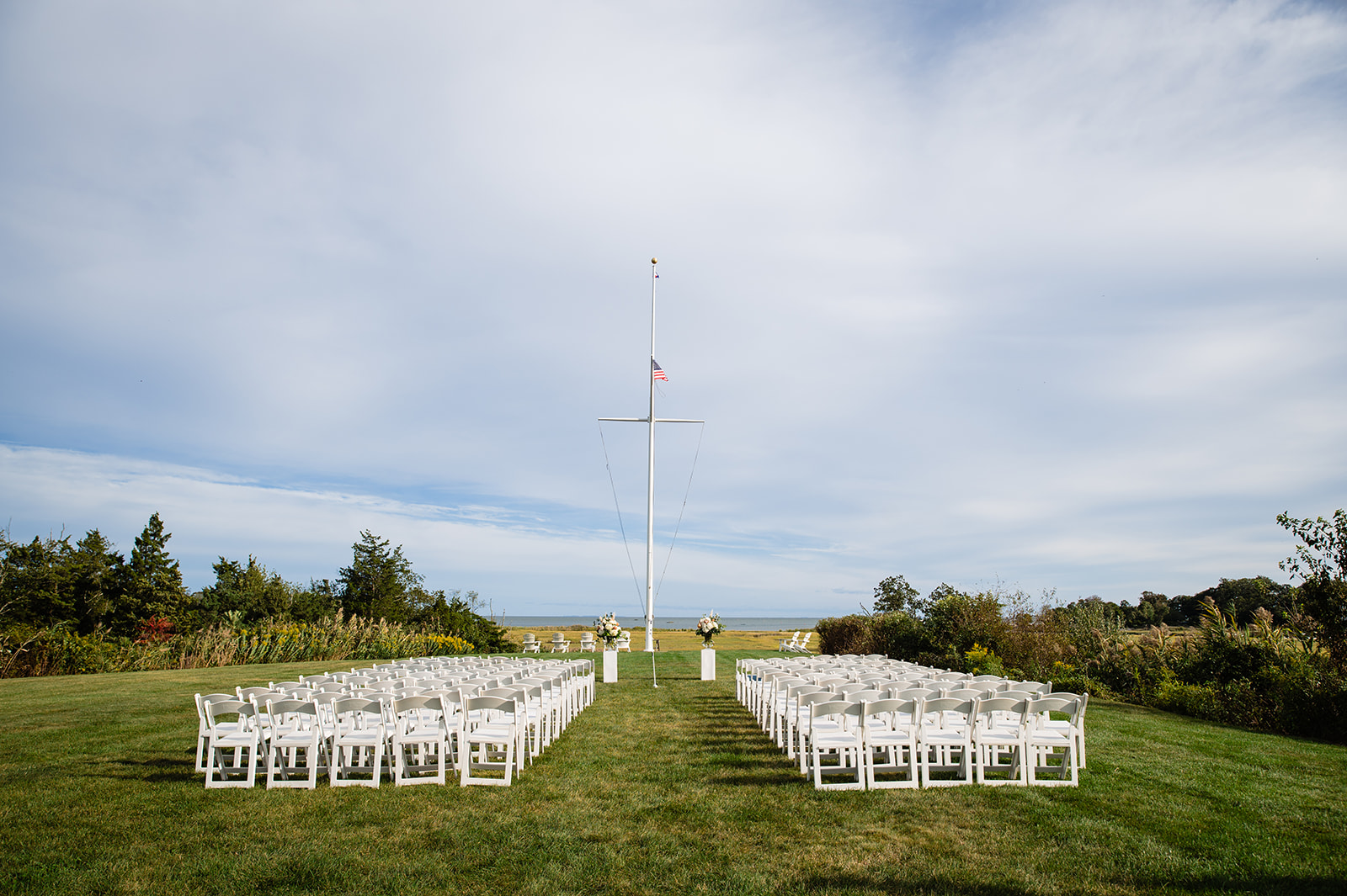 A serene outdoor wedding setup with rows of white chairs facing a flagpole on a lush green lawn, a clear blue sky above, and the ocean in the distance.