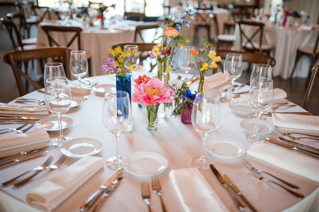 Elegantly set wedding reception table with a white linen tablecloth, crystal glassware, and vibrant centerpieces of wildflowers in assorted colored glass vases.