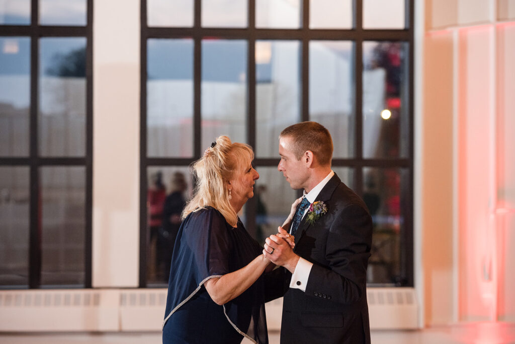 Mother and son sharing a touching dance moment at a wedding reception, with large windows in the background illuminating the space with natural light.