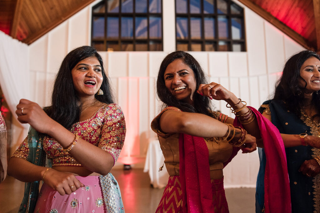 Group of women in colorful traditional Indian attire dancing and laughing joyously at a wedding celebration.