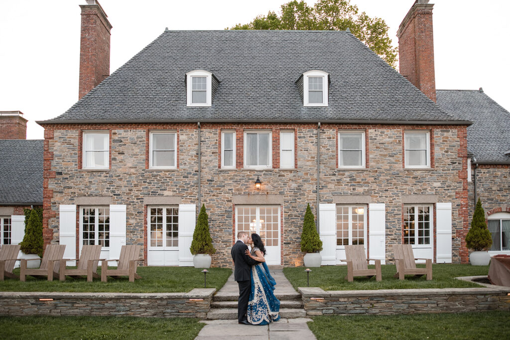 Bride and groom embracing outside a stately brick manor with white doors and windows, under a clear sky at dusk.