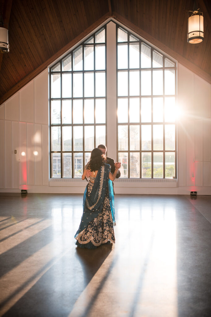 Bride and groom sharing a dance in a bright hall with tall windows, with the sun setting in the background casting a warm glow.