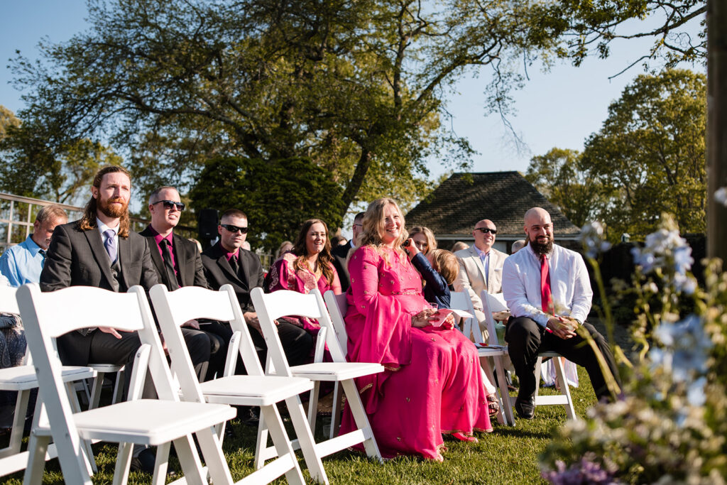 Wedding guests seated in white chairs during an outdoor ceremony with a backdrop of large trees and a clear blue sky.