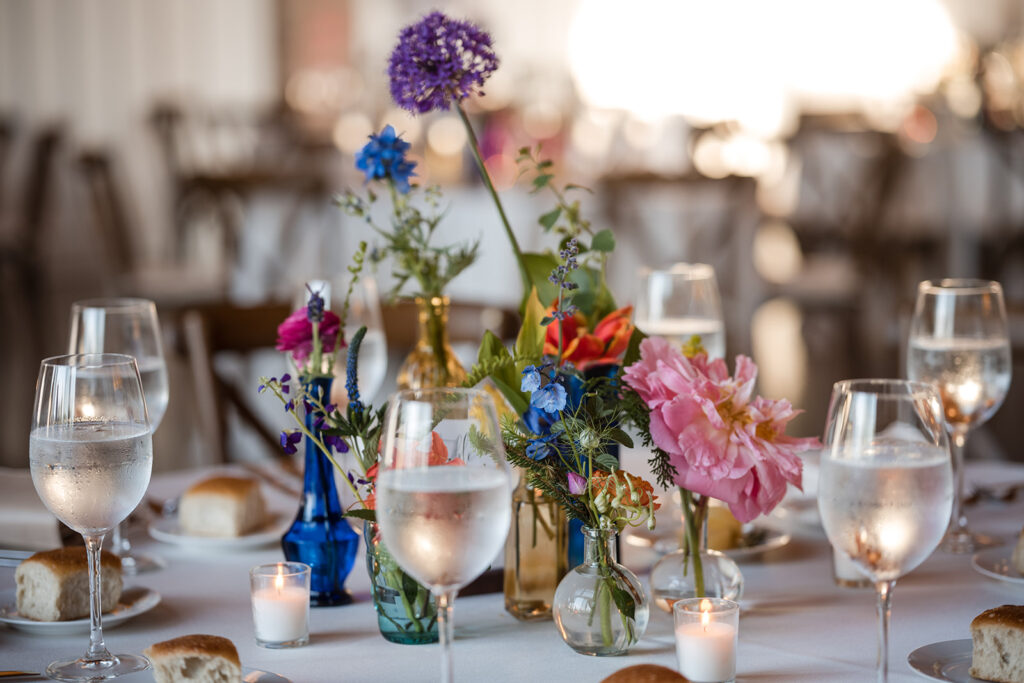 Close-up of a wedding table setting, featuring delicate glassware, white plates, and a colorful flower arrangement amidst the golden hour light.
