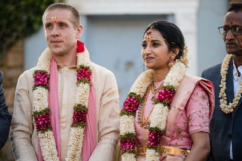 A bride and groom in traditional Indian wedding attire, the groom in a cream sherwani with a pink stole and the bride in a pink saree, both adorned with elaborate flower garlands.
