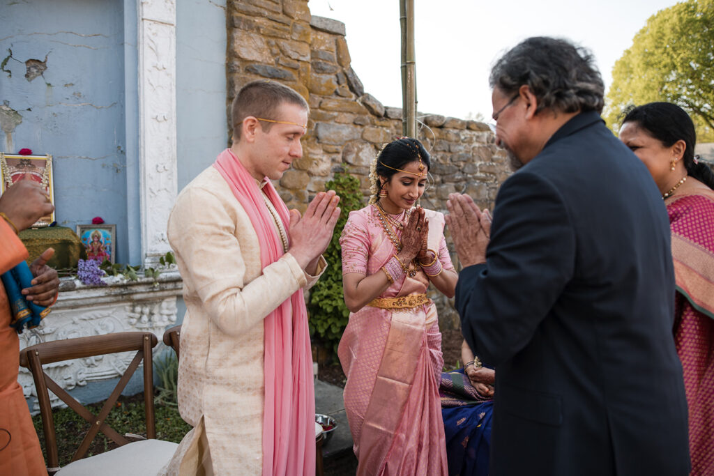 The bride and groom performing a traditional Indian wedding ritual, surrounded by family, with the groom in a pink stole and the bride in a pink saree, both with hands clasped in a gesture of prayer.