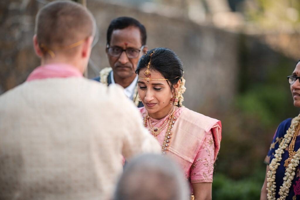 The bride, dressed in a pink saree and adorned with gold jewelry, looks down thoughtfully during a wedding ceremony with family members surrounding her.