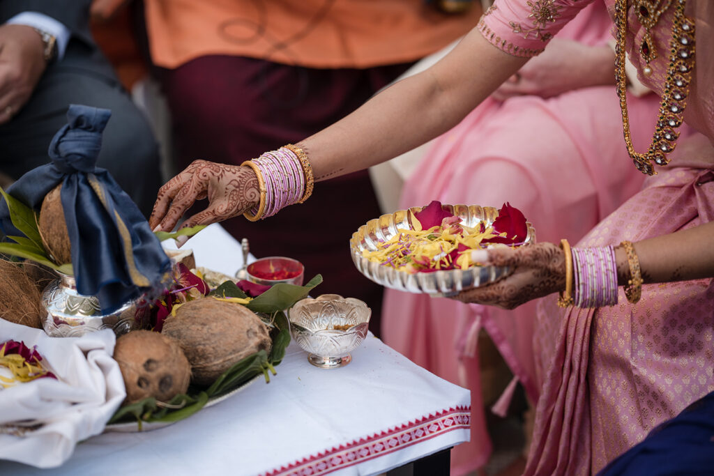 A traditional Indian wedding ritual with the bride in a pink saree reaching out to a ritualistic silver plate, hands adorned with henna and bangles.