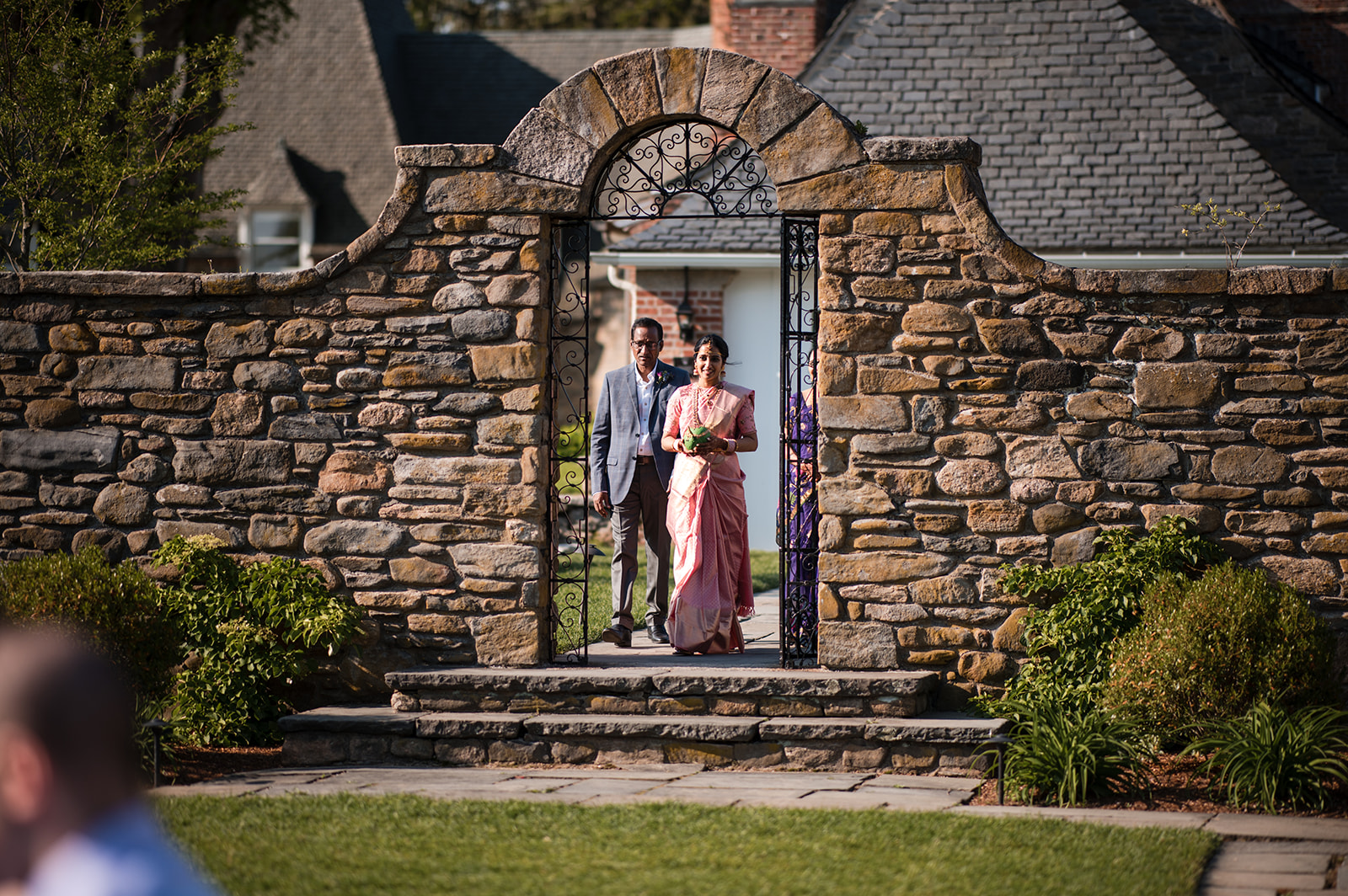 The bride and groom walking through a stone archway in a garden, the bride in a pink saree holding a bouquet, and the groom in traditional attire.