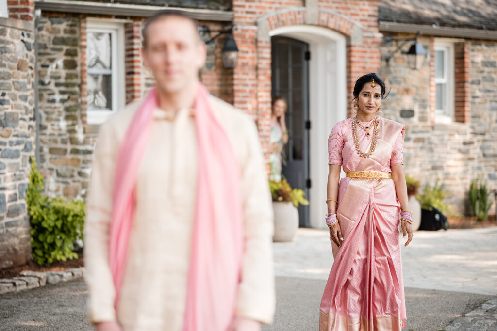 The bride in a pink saree, adorned with gold jewelry, walking towards the camera with a focused expression, while the groom stands in the background out of focus.